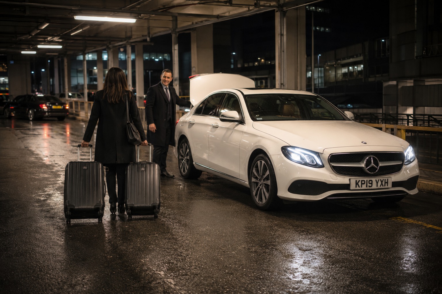 Traveller wheeling suitcases to a waiting saloon taxi at a Manchester Airport pick-up zone at night